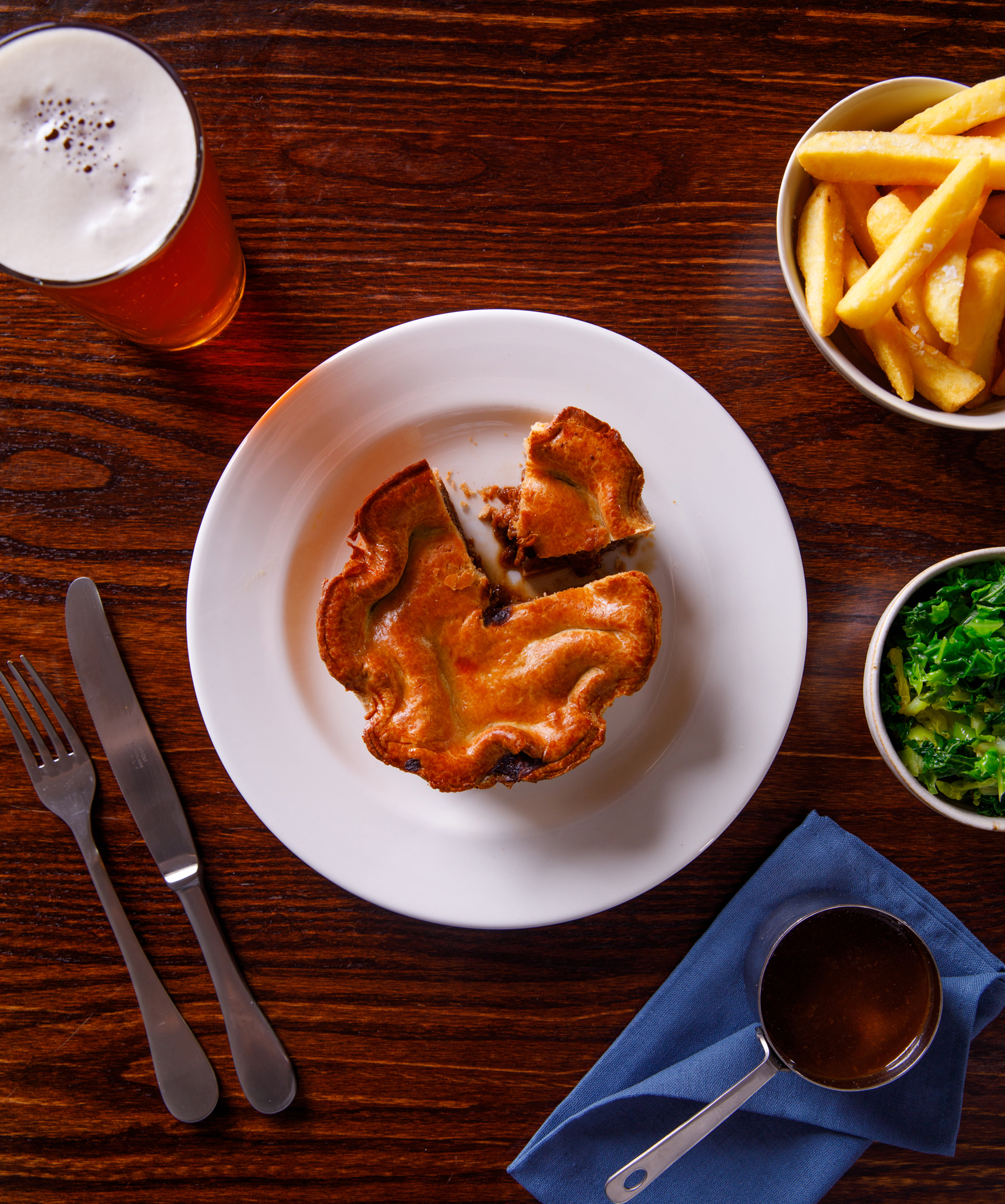 photo from above of a steak and ale pie. Pie is surrounded by a pint of beer, a bowl of chips, greens and a small pan of gravy