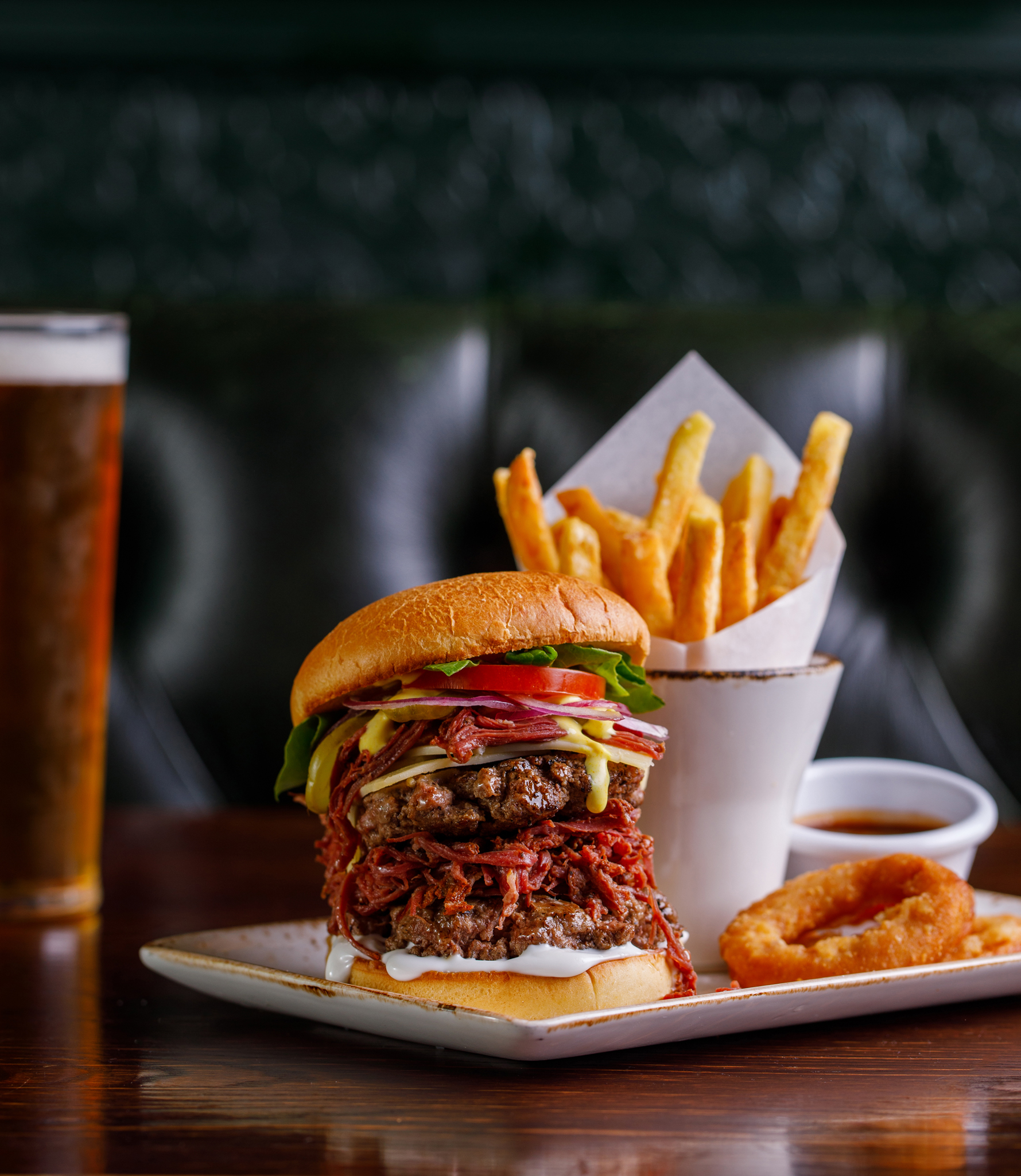 photo of a big burger with pulled pork and drippy sauce. Chips behind the burger and onion rings on the side