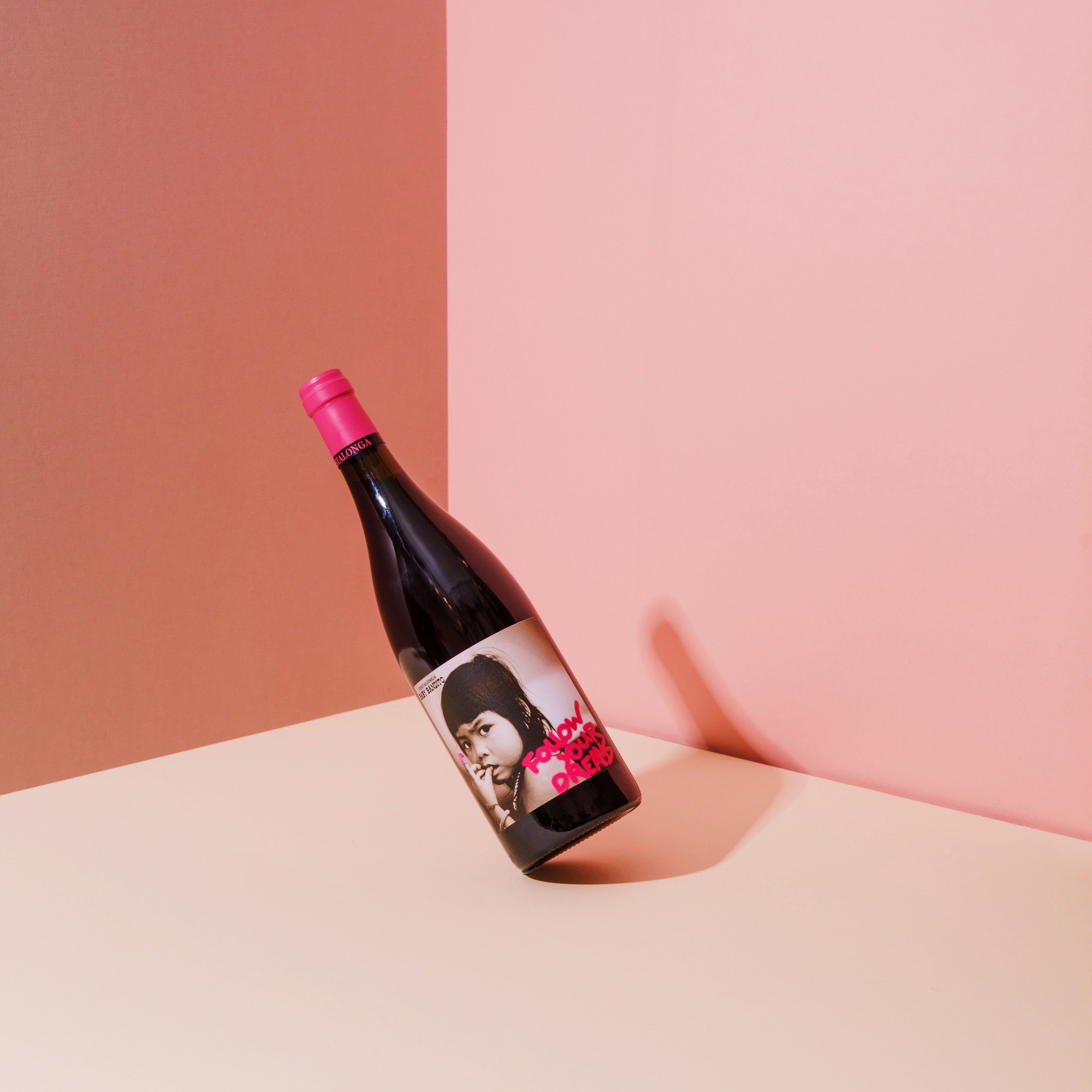 Bottle of wine balanced on edge leaning backwards unnaturally. Background colours pink and mauve