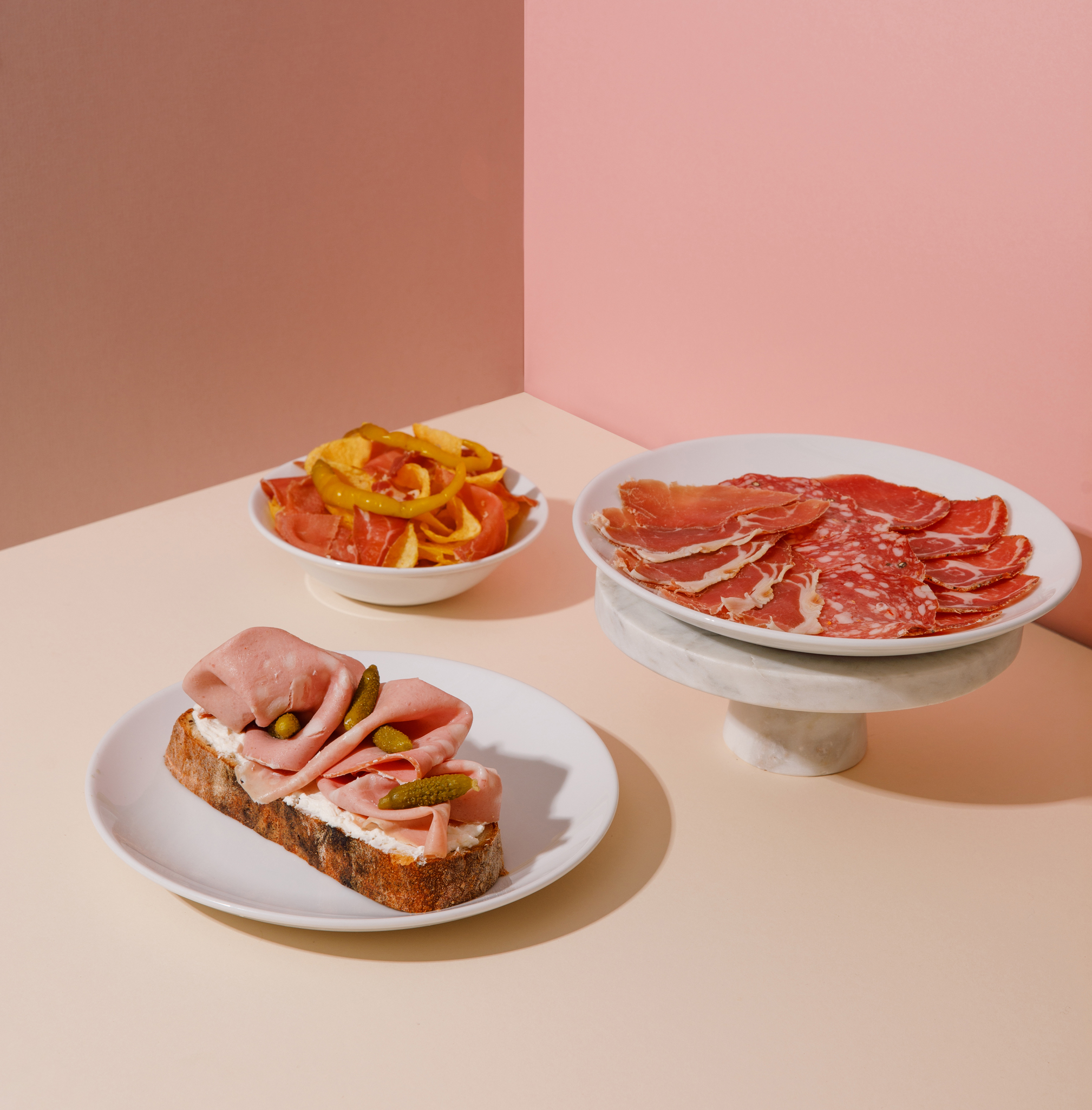 Scene of 3 plates of food on pink and mauve background. Plate of cured meats, a bowl of crisps and cured meats and a plate with mortadella on sourdough