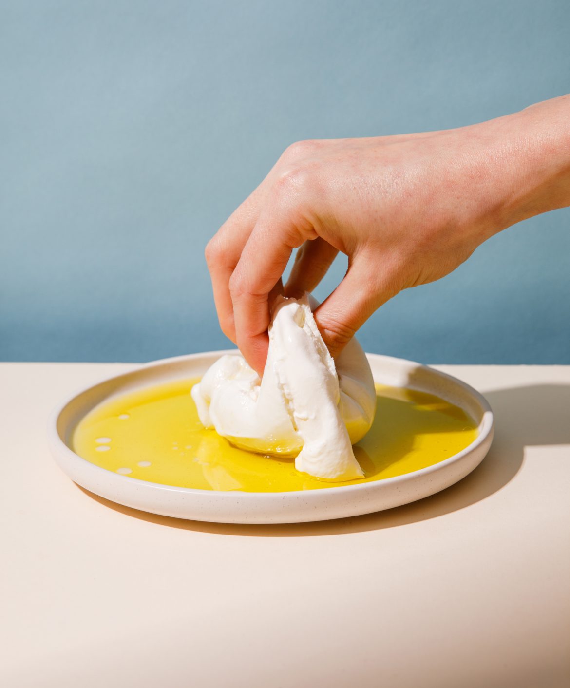 photograph of a hand grabbing a fresh burrata on a plate. Background is pale blue, hand is coming from right to left