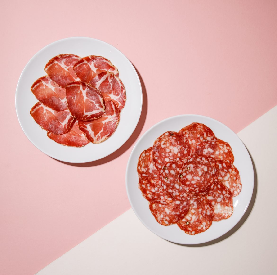 photograph of two plates of cured meats from above. One plate is gabagool, one plate is salami
