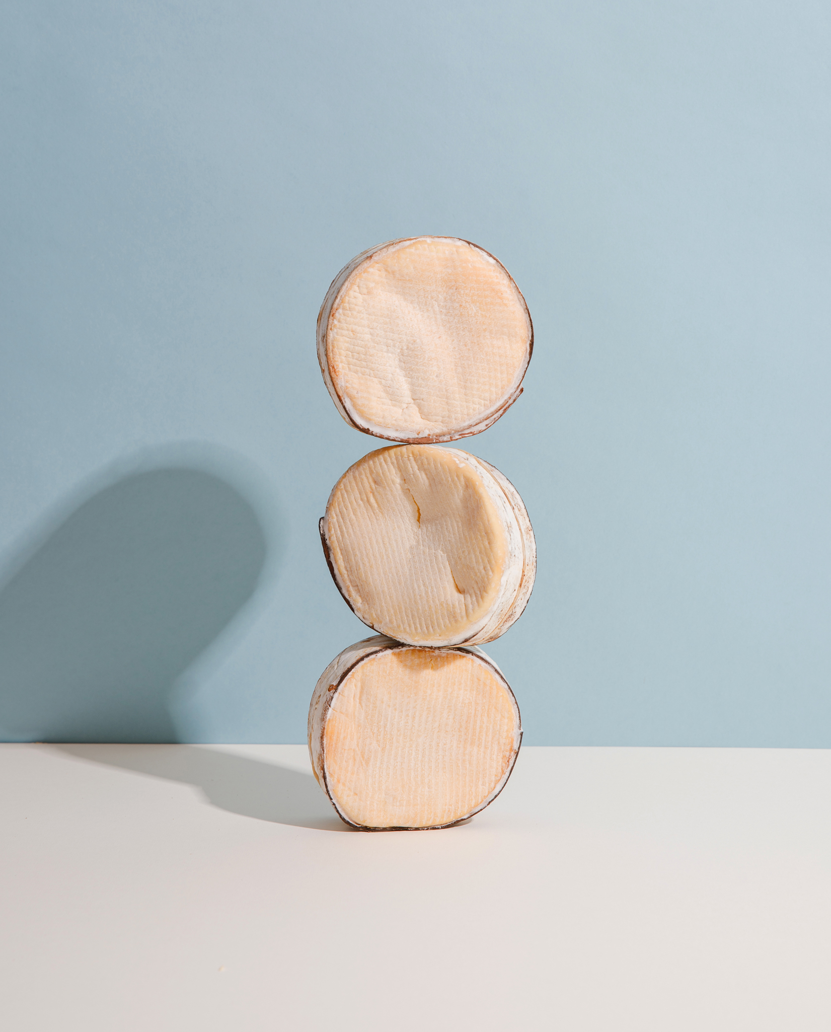 photograph of three small round cheeses stacked on top of each other end to end. Background is blue and surface is off white