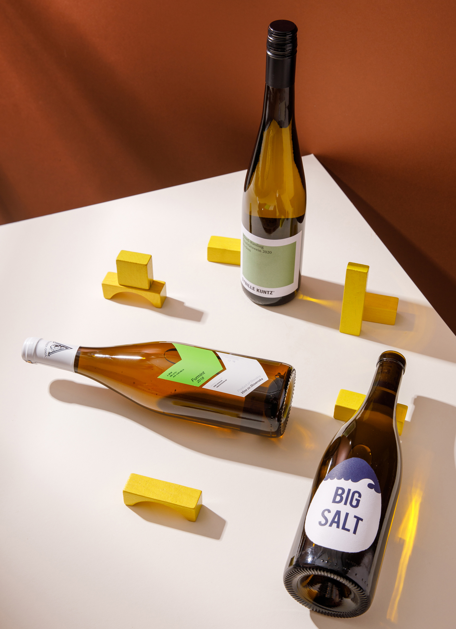 photograph of three bottles of white wine arranged geometrically with yellow wooden blocks filling the spaces on the table. Table is off white in colour, background is chestnut brown