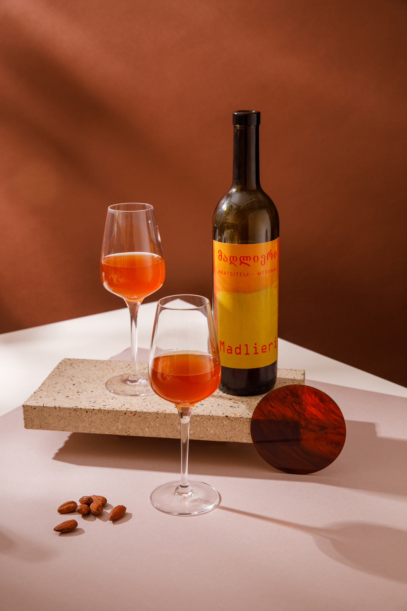 photograph of a bottle of orange with with two glasses of orange wine next to it. One glass and the bottle stand on a small concrete plinth, background is chestnut brown