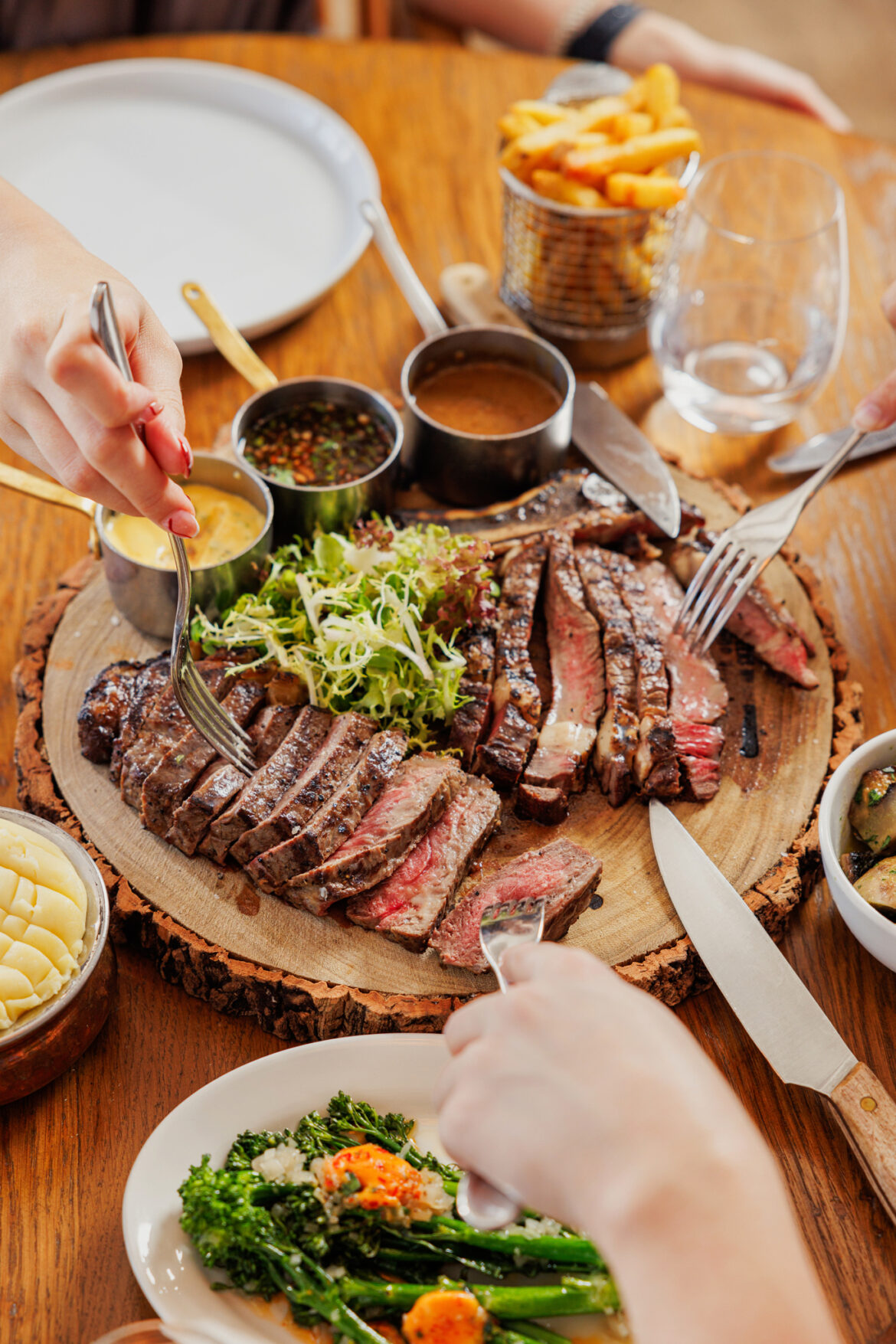 A photo of a steak on a wooden board being shared by three people. The steak is cooked medium rare and there are sides of mashed potato and broccoli.