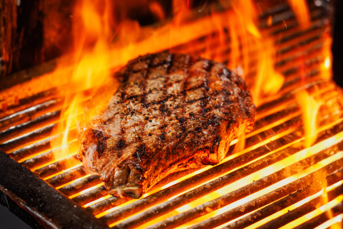 A photo of a steak on a char grill surrounded by flames