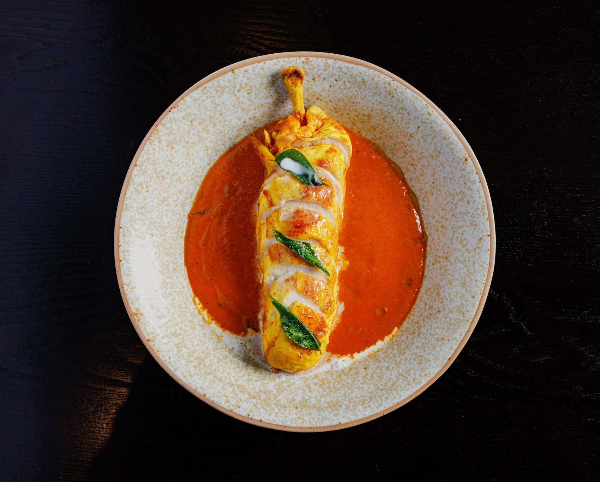 Stuffed chicken leg curry at Pravaas — Close-up of a stuffed chicken drumstick in a rich red curry sauce, photographed against a dark background.