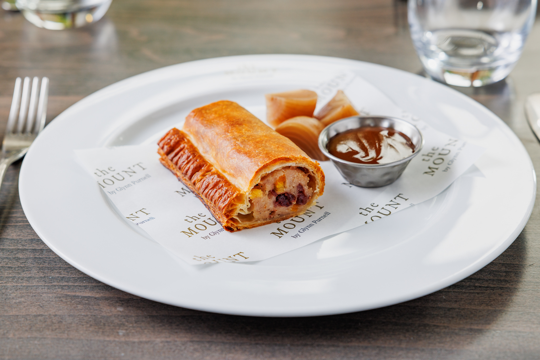 Gourmet sausage roll served with pickled vegetables and a pot of brown sauce on branded paper at The Mount by Glynn Purnell.