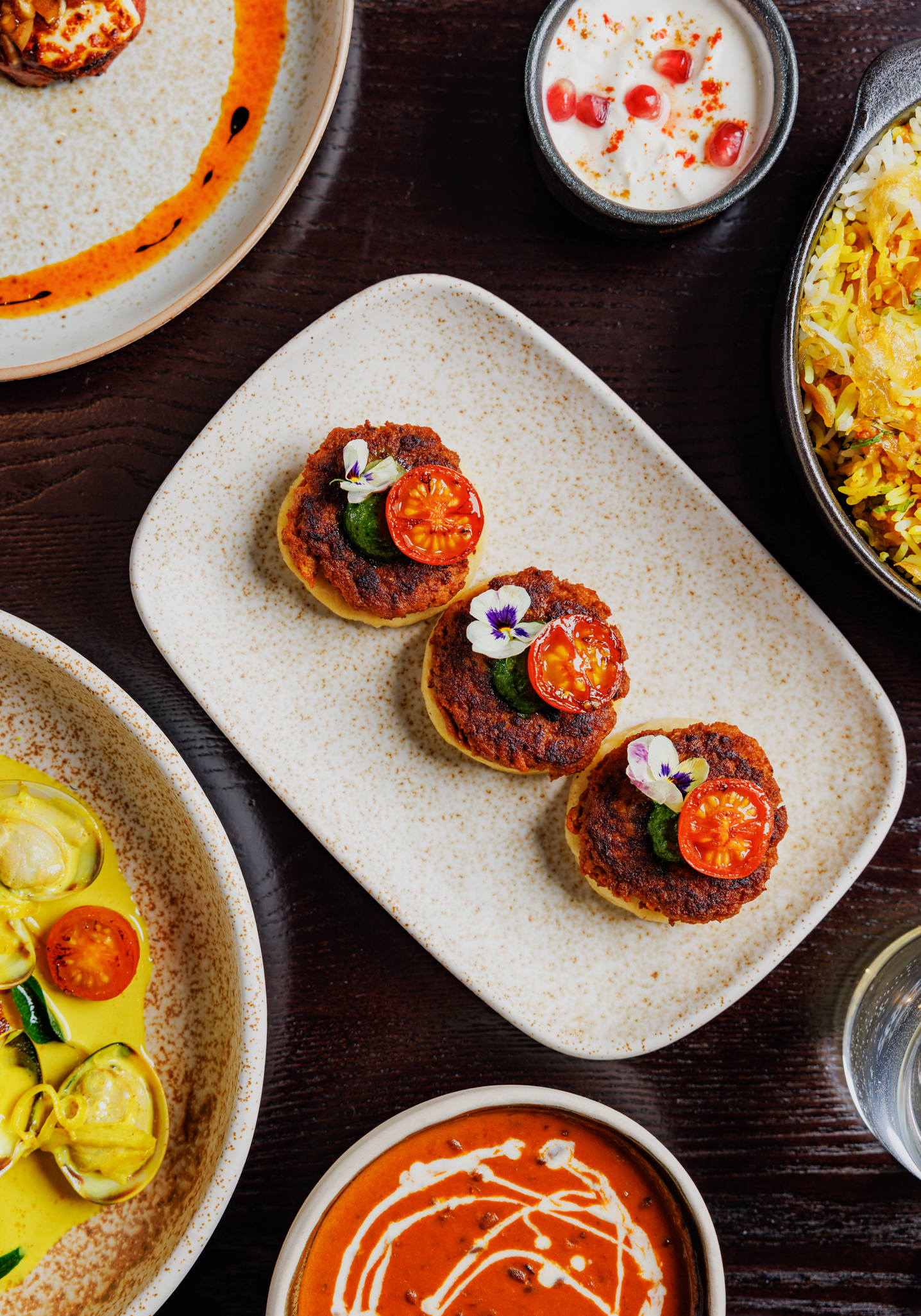 Close-up of three lamb galouti kebabs topped with cherry tomatoes and edible flowers, surrounded by vibrant Indian dishes.