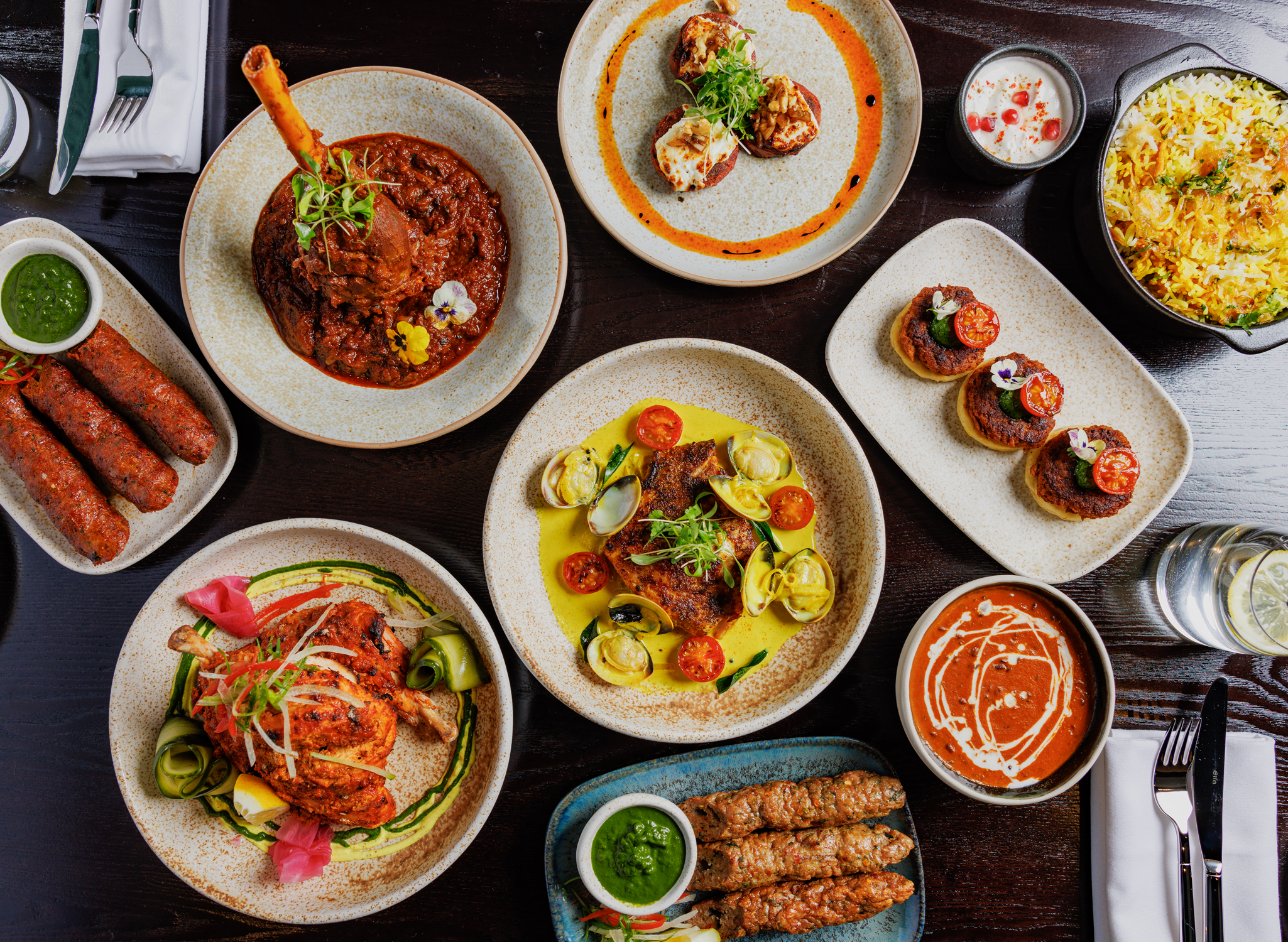 Full overhead table shot of colourful Indian dishes at Pravaas, featuring lamb shank curry, biryani, kebabs, and fish curry.